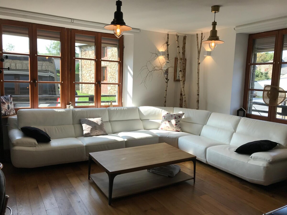 A spacious living area features a white sectional sofa, complemented by a wooden coffee table. Large windows provide natural light, with a view of the surrounding nature. Industrial-style light fixtures hang elegantly from the ceiling, creating a warm ambiance in the room.