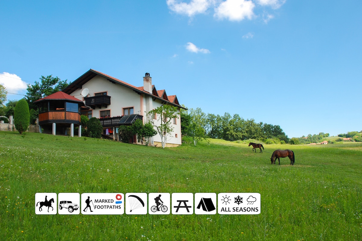 A spacious eco-farm building is visible amidst a lush green field, surrounded by trees. Two horses are grazing peacefully in the foreground under a clear blue sky. Symbols for outdoor activities such as horseback riding, hiking, and cycling are displayed at the bottom.