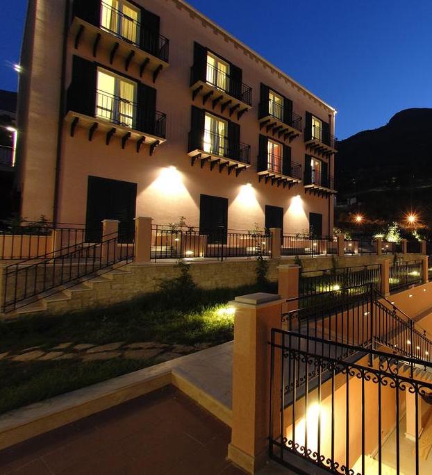 The exterior of a three-story building is showcased, featuring a pink façade with dark window shutters and balconies. Soft lighting illuminates the stairs and surrounding garden, creating a welcoming ambience. A backdrop of mountains is visible, enhancing the serene setting.