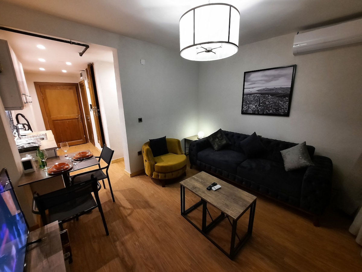 A living area features a dark sofa accompanied by decorative pillows and a cozy yellow armchair. A wooden coffee table sits in front, and a dining area with a table for four is present. Soft lighting emanates from a stylish ceiling fixture.