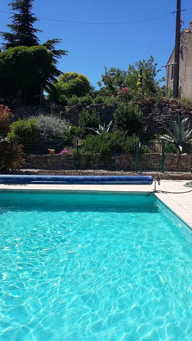 A heated swimming pool is visible, featuring clear blue water reflecting the sunlight. Surrounding greenery includes various shrubs and plants, creating a serene setting. The pool area is bordered by a tiled surface and a low stone wall, enhancing the outdoor experience.