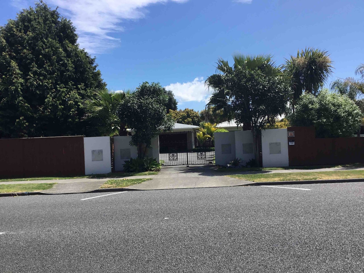 The exterior of a secure property is framed by tall palm trees and green shrubs. A gated entrance is visible amidst a well-maintained garden, creating a private atmosphere. The clean driveway leads to the entrance, with clear skies above and a smooth road in front.
