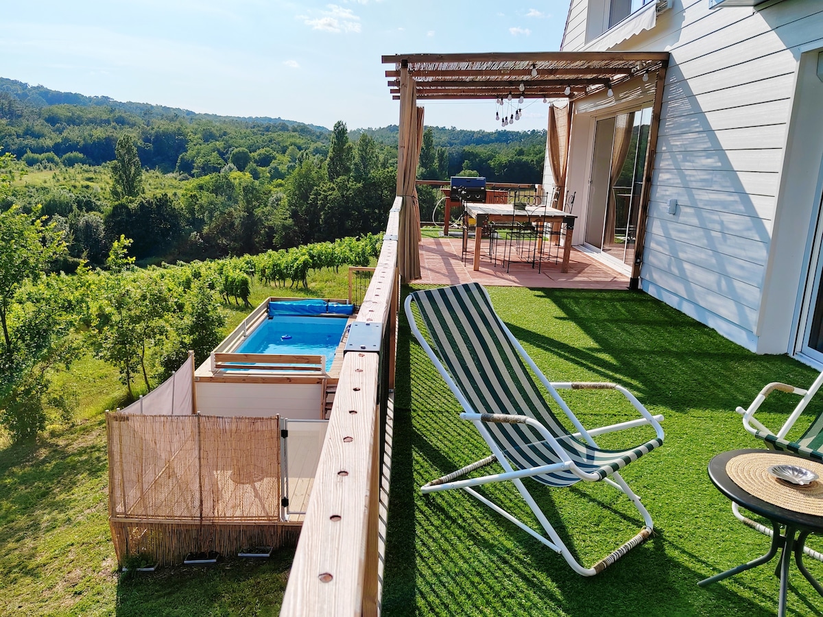 An elevated terrace features outdoor dining furniture and a shaded pergola, overlooking a lush landscape. A small, above-ground pool is visible below, surrounded by greenery. Loungers are positioned on the terrace, providing a relaxing space to enjoy the scenic view.