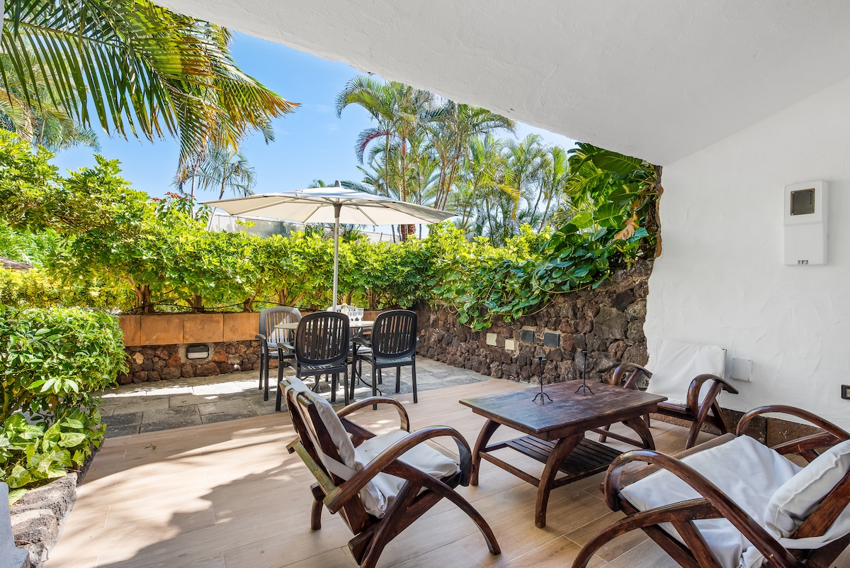 A private terrace features a wooden table surrounded by four black chairs, complemented by two reclining armchairs. Lush greenery provides a natural backdrop, and a large umbrella offers shade, creating a serene outdoor space for relaxation and dining.