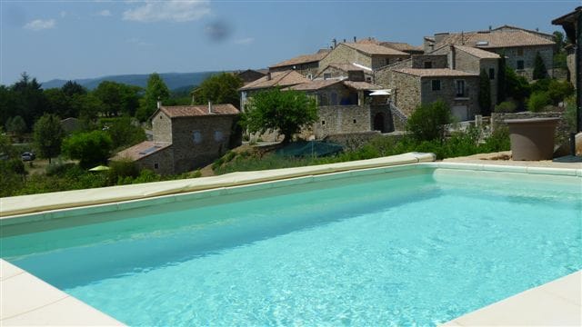 A refreshing swimming pool is set against a backdrop of rustic stone buildings. The clear water reflects the sky, while the surrounding landscape features gentle hills and greenery. The serene environment invites relaxation and leisurely afternoons.