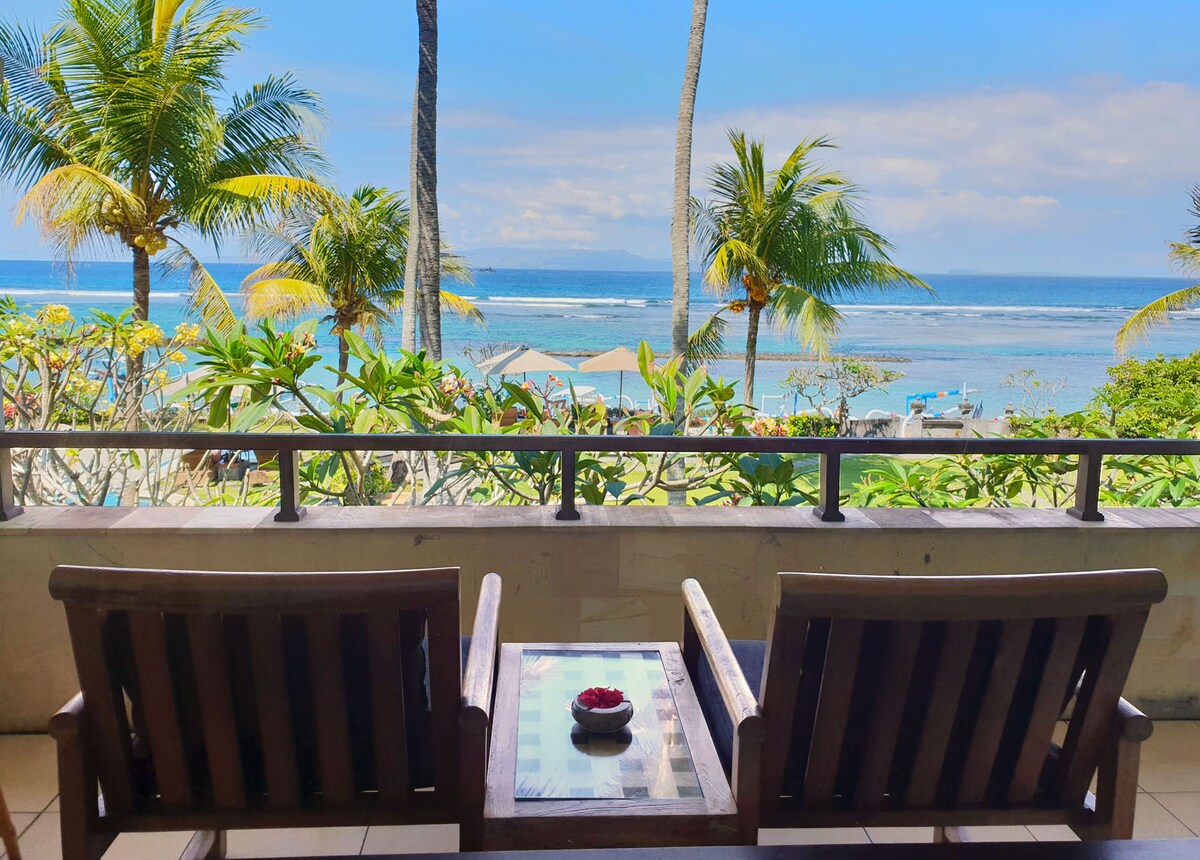 Two wooden chairs are positioned on a private balcony, facing a view of the tranquil sea and palm trees. A small table holds a decorative bowl with flowers. The open sky and gentle waves create a serene beach atmosphere.