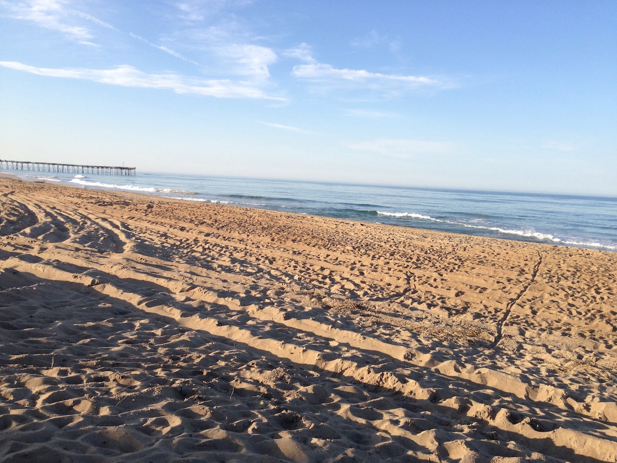 A serene beach landscape features smooth, sandy shores stretching towards gentle waves. A long pier extends into the calm ocean, under a clear blue sky with scattered clouds. The tranquil scene invites relaxation and connection with nature.
