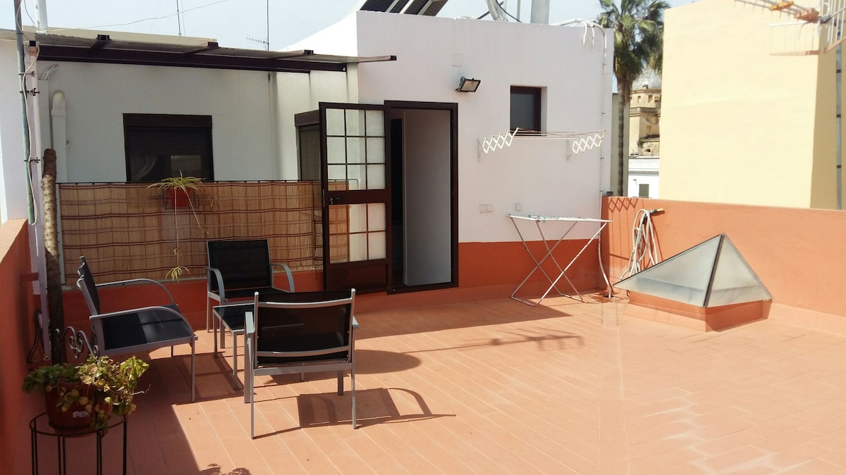 A spacious terrace is presented, featuring several outdoor chairs and a small table. Sunlight brightly illuminates the area, which is surrounded by low walls. A laundry drying rack stands against the wall, and a glass structure is visible in the corner.