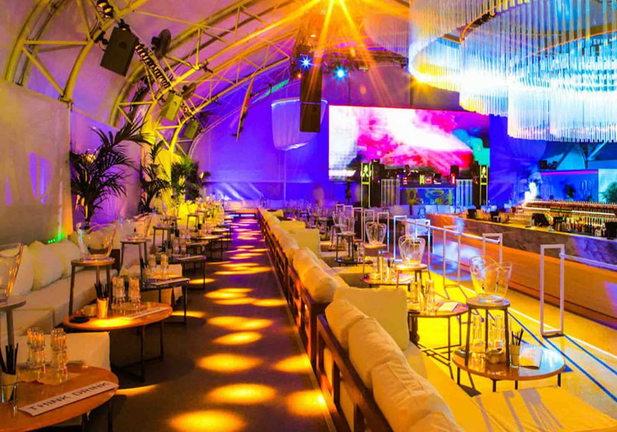 An expansive indoor space features a stylish lounge area with multiple seating arrangements around small tables. A vibrant light display bathes the room in colors, while a large screen displays visuals in the background. Clear glass lighting fixtures hang overhead, adding a modern touch.