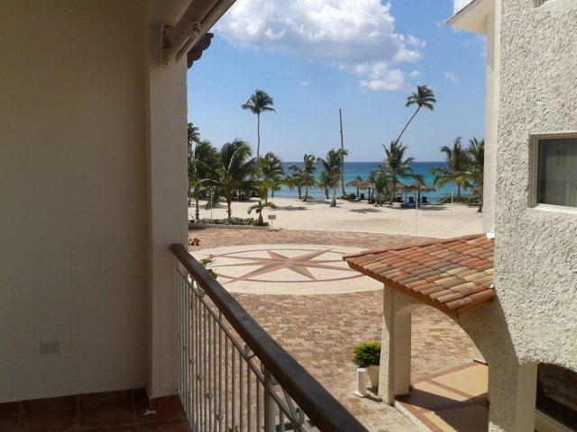 A view from the balcony reveals a sandy beach lined with palm trees, leading to clear turquoise waters. The pathway features circular stone patterns, and guests can see the inviting beach area in the distance with a blue sky dotted with fluffy clouds.