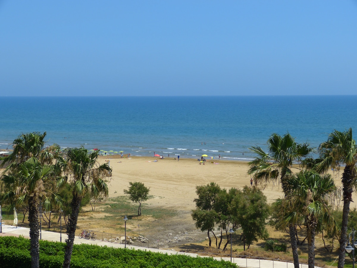 A view of the sandy beach stretches towards the calm sea, framed by palm trees in the foreground. Sunbathers and umbrellas dot the shoreline, with a clear blue sky above, creating a serene coastal setting.
