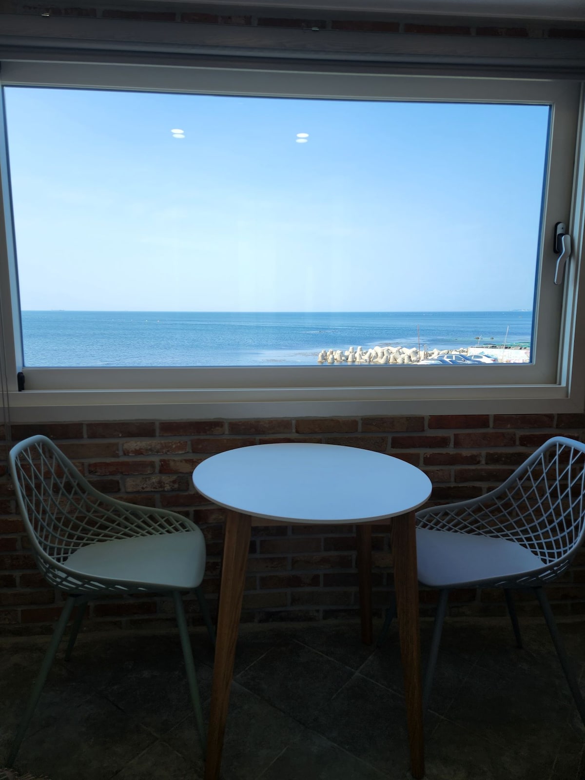 A small round table is positioned near a large window, offering a view of the serene sea. Two modern chairs, one white and one light green, frame the table. The shoreline is visible in the distance, complemented by a clear blue sky.