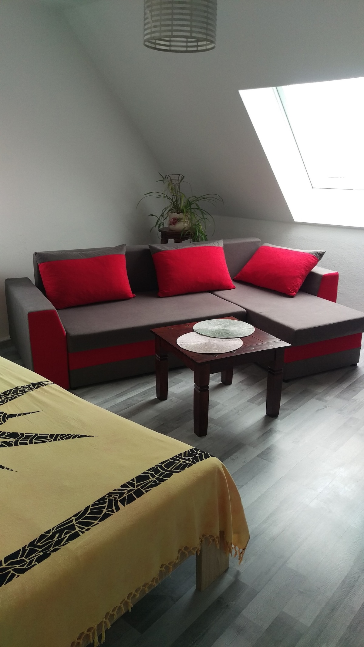 A comfortable living area is depicted, featuring a gray and red sofa accented with vibrant cushions. A small wooden table provides a surface for items. Natural light enters through a large window, illuminating the space and highlighting the soft grey flooring.