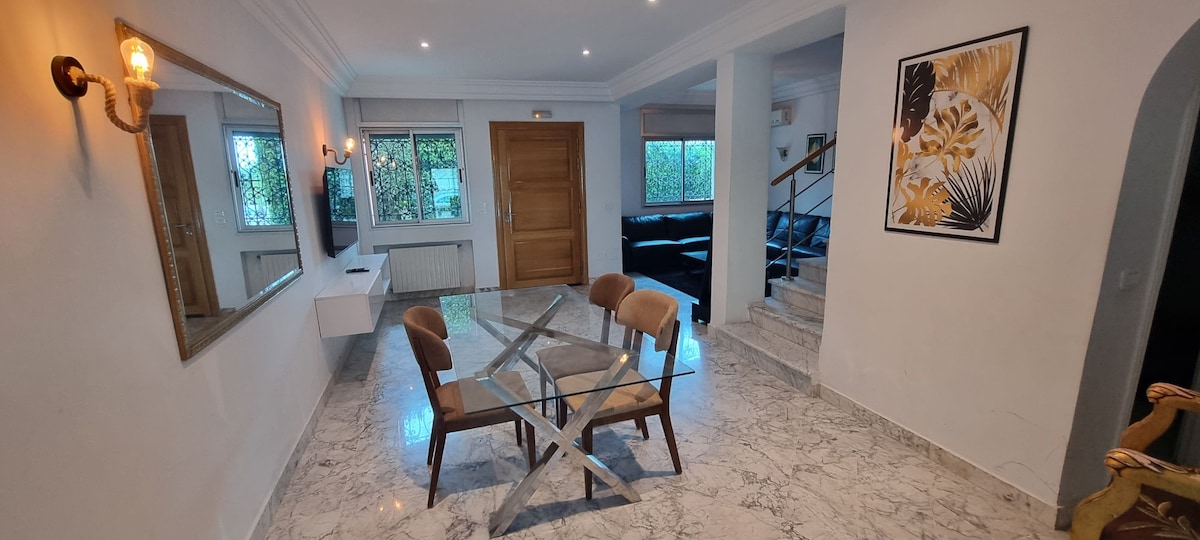 A spacious dining area features a glass table surrounded by two wooden chairs. Natural light is allowed to flow through large windows, while a cozy seating arrangement is visible in the background. A decorative mirror enhances the sense of openness in the room.