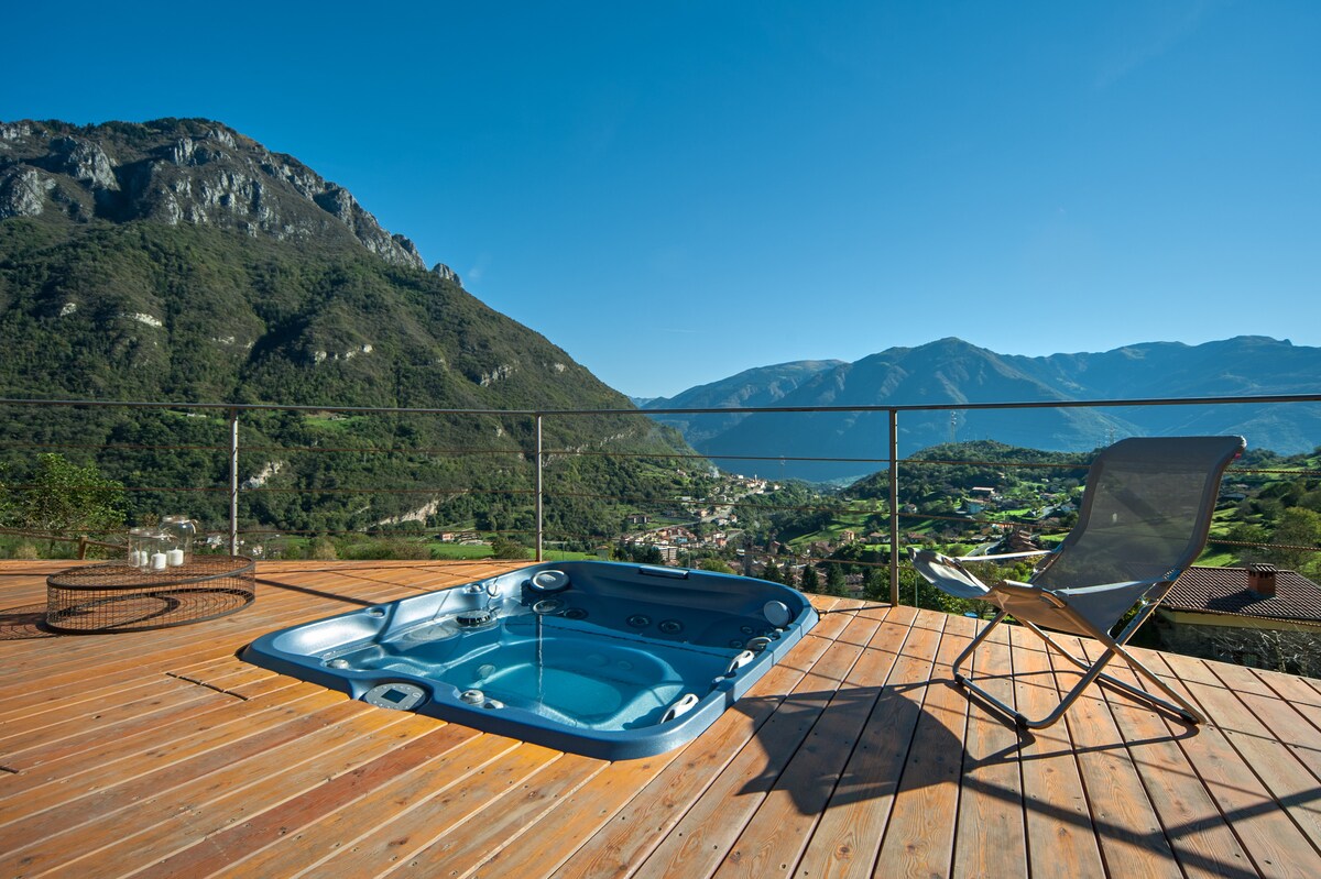 A spacious terrace reveals a hot tub alongside a reclining chair, framed by sweeping views of the valley and mountains. Natural wooden decking enhances the outdoor setting, while a clear sky provides an inviting atmosphere for relaxation.