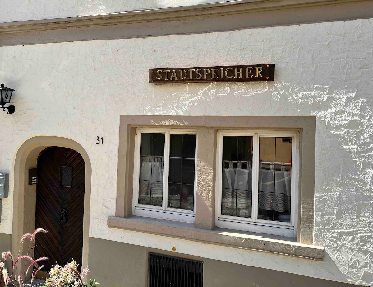 The building's exterior features a light-colored facade with a textured finish. A wooden sign labeled 'STADTSPEICHER' is mounted above two windows, which are adorned with simple curtains. The entrance is framed by a curved archway, adding architectural character.