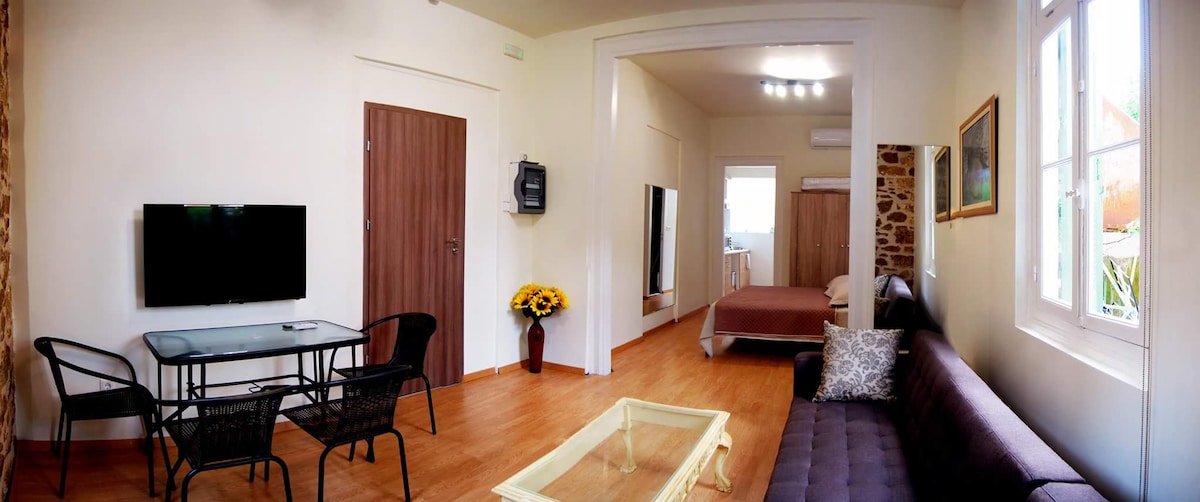 A well-lit living area includes a cozy sofa, a glass coffee table, and a dining table with four black chairs. A television is mounted on the wall opposite the sofa. Natural light filters in through the nearby window, highlighting the warm wood floor.