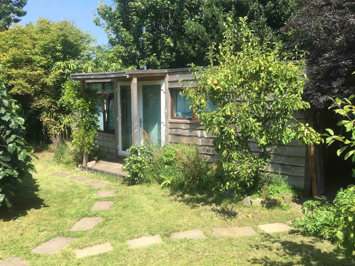 A self-contained garden cabin is nestled among greenery, featuring large windows that invite natural light. A stone pathway leads to the cabin entrance, surrounded by various plants and trees, creating a peaceful outdoor setting.