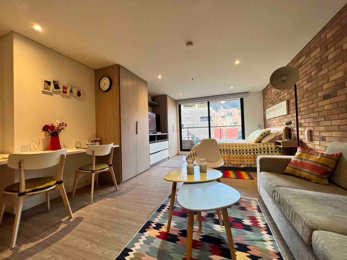 An open layout showcases a cozy living area furnished with a sofa and two coffee tables. A dining space with white chairs sits next to a modern kitchen. Light wood cabinets and exposed brick walls add texture, while large windows offer views of the outdoor scenery.
