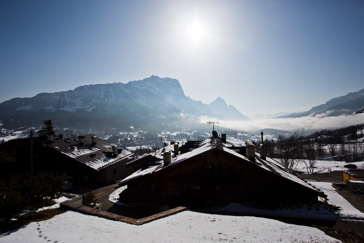 A winter landscape is displayed, featuring snow-covered rooftops and distant mountains. The sun casts a bright light over the scene, highlighting the natural beauty of the area. Soft clouds linger in the valley below, enhancing the tranquil ambiance.