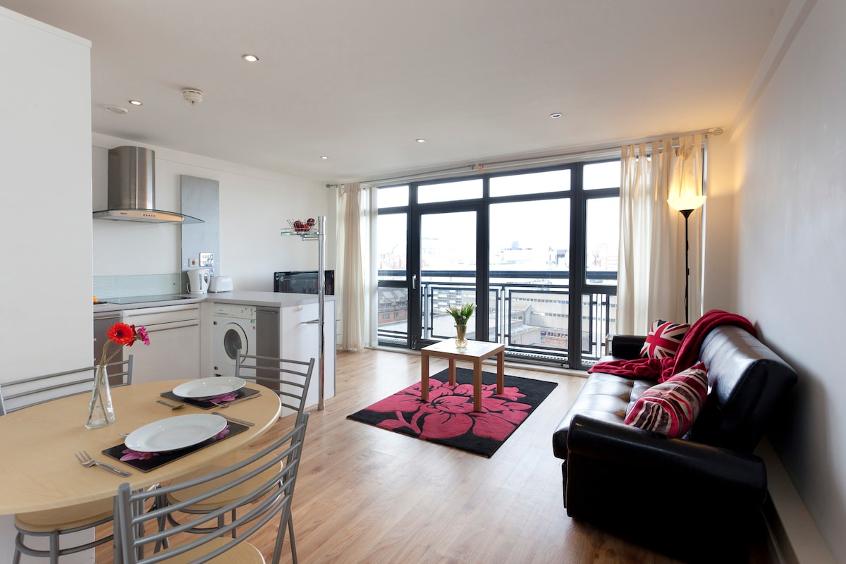 A bright living space features large windows allowing ample natural light. A modern kitchen is seen alongside a round dining table with place settings. A black sofa and an area rug with bold floral patterns create a cozy seating area. A washing machine is included in the layout.