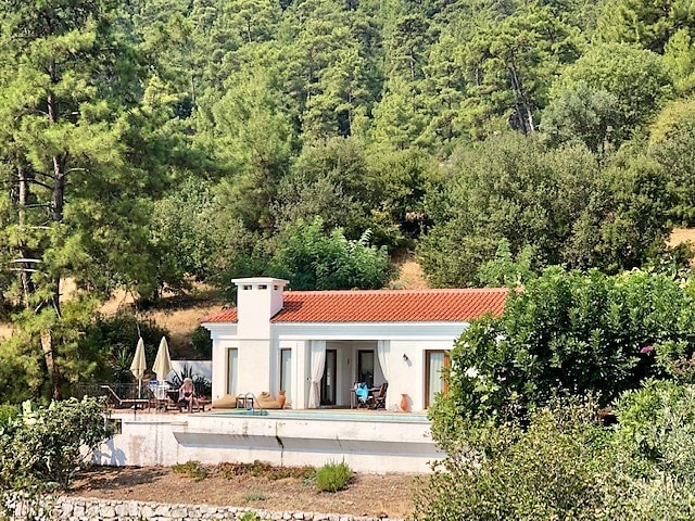 A villa with a red-tiled roof is nestled among lush greenery. It features large glass doors that open to a patio, which is equipped with outdoor furniture. Two sun umbrellas provide shade, while surrounding trees enhance the serene atmosphere.