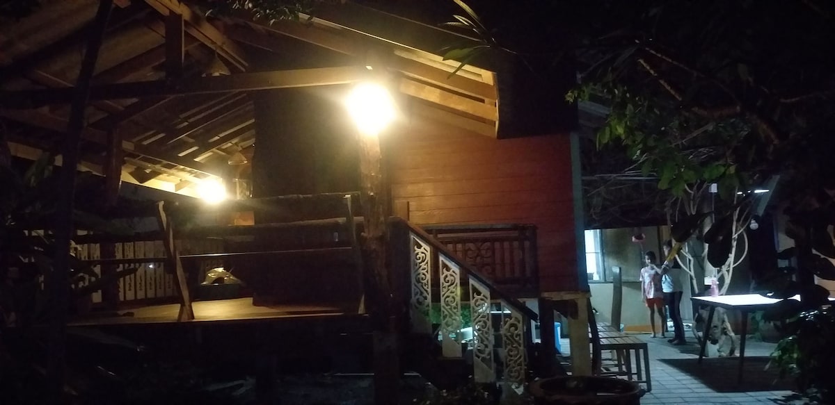 A wooden structure is illuminated by warm lights, showcasing a spacious porch and intricate railing patterns. Cozy seating areas are presented on the outdoor deck, surrounded by lush greenery. The nearby path leads towards additional living spaces, enhancing a sense of privacy.