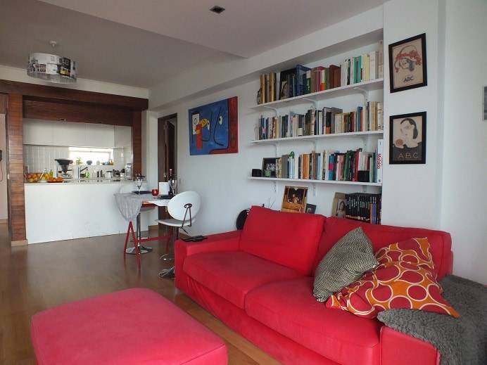 A bright and modern living area is complemented by a red sectional sofa adorned with patterned cushions. A small ottoman sits nearby. A wall-mounted bookshelf displays a variety of books, while a colorful artwork adds a pop of interest above the sofa. The open kitchen is visible in the background.