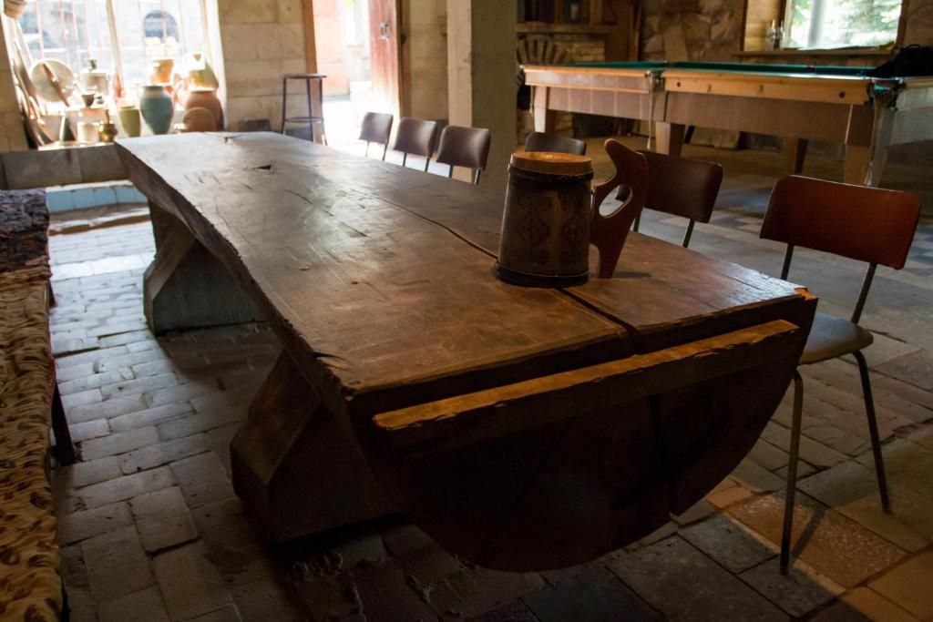 A rustic wooden table occupies the center of the space, featuring a unique design with a smooth surface and pronounced edges. Surrounding the table are several simple chairs. The setting is illuminated by soft natural light filtering through nearby windows.
