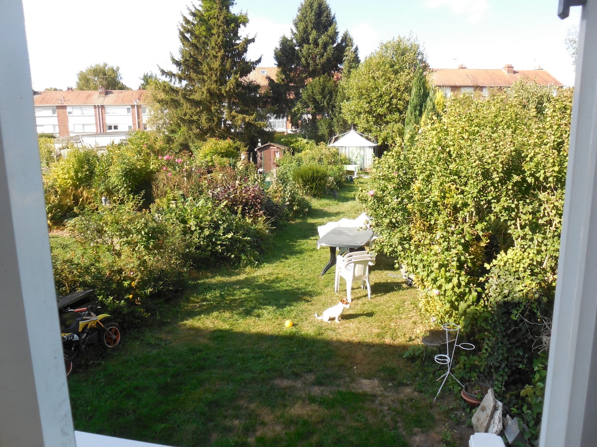 A spacious garden area is depicted, featuring diverse greenery and colorful plants. A white table and chairs are positioned among the foliage, offering a relaxing spot for outdoor enjoyment. A gazebo is partially visible, along with a small shed, creating a natural, inviting environment.