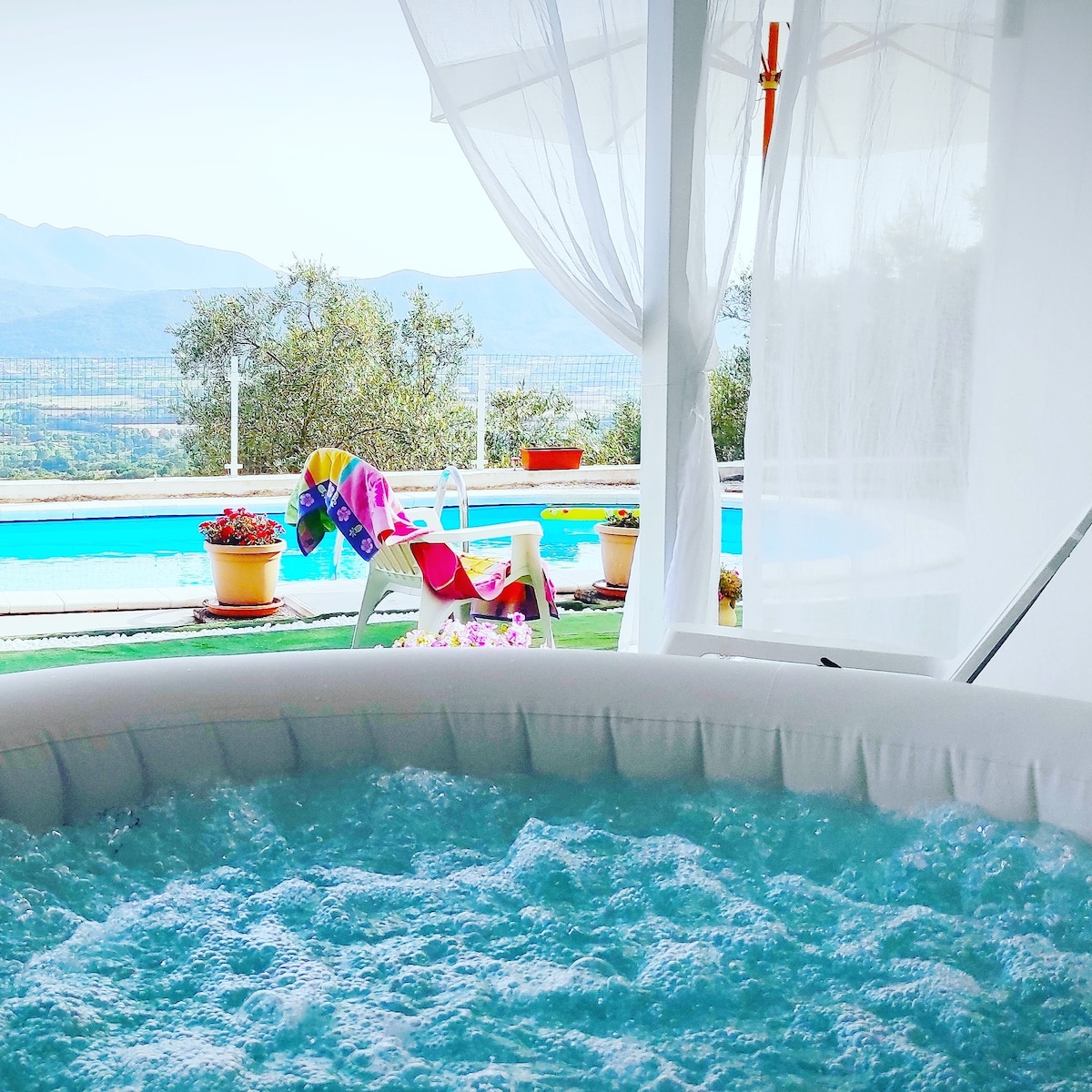A bubbling hot tub is visible in the foreground, surrounded by transparent curtains. In the background, a swimming pool is partially shown with lounge chairs and colorful towels nearby, framed by scenic views of rolling hills and olive trees.