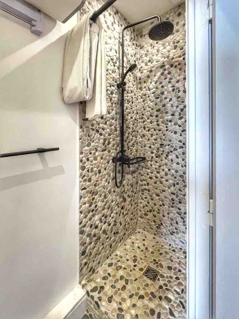 The shower area features a stone pebble texture on the walls and flooring, enhancing the natural appearance. A rainfall showerhead is mounted above, with a wall-mounted holder for towels nearby. A small shelf is provided for toiletries.