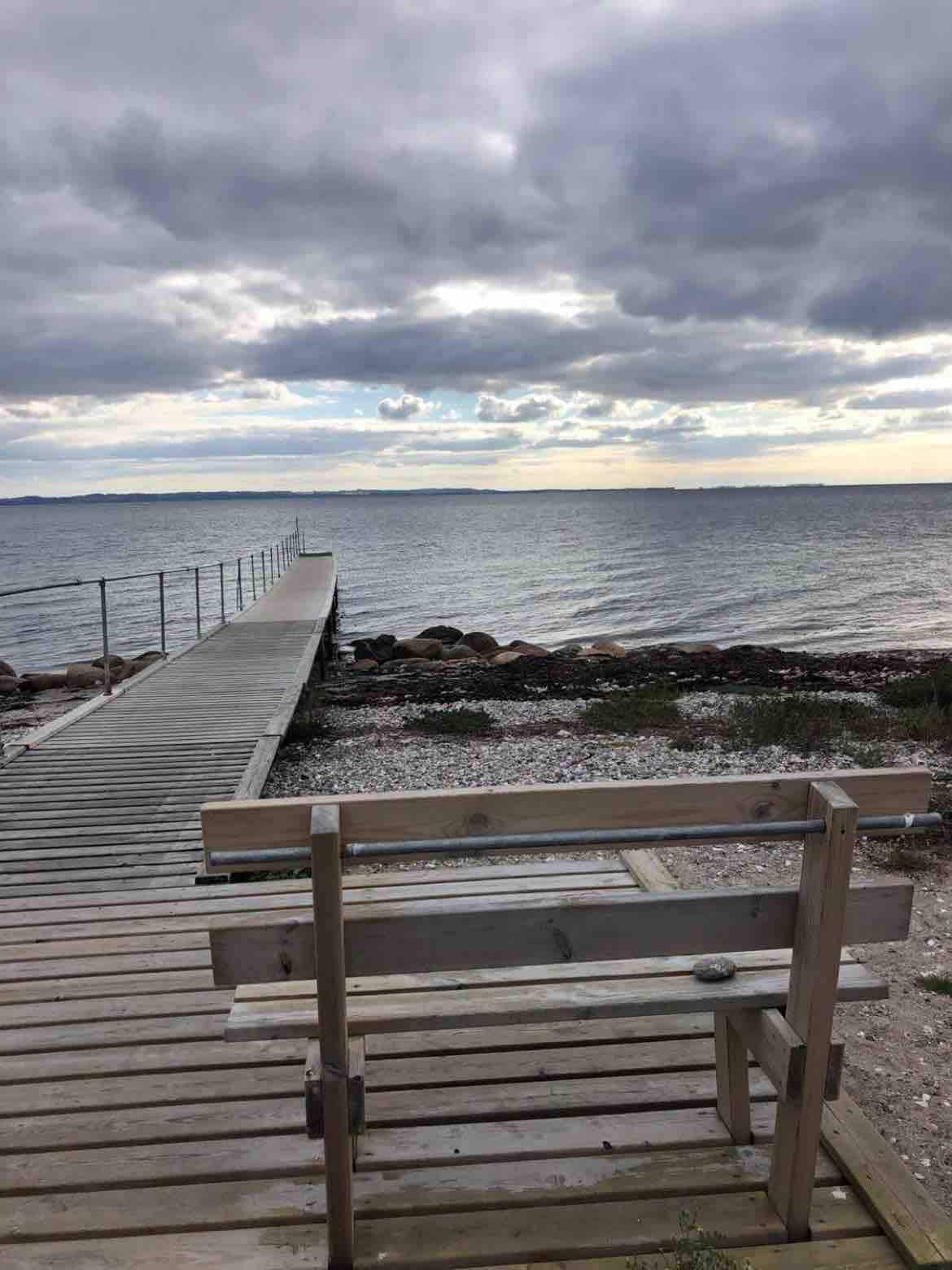 A wooden walkway extends into the water, bordered by pebbles and plants. A simple wooden bench is positioned nearby, offering a view of the calm sea under a cloudy sky with varying shades of gray and blue.