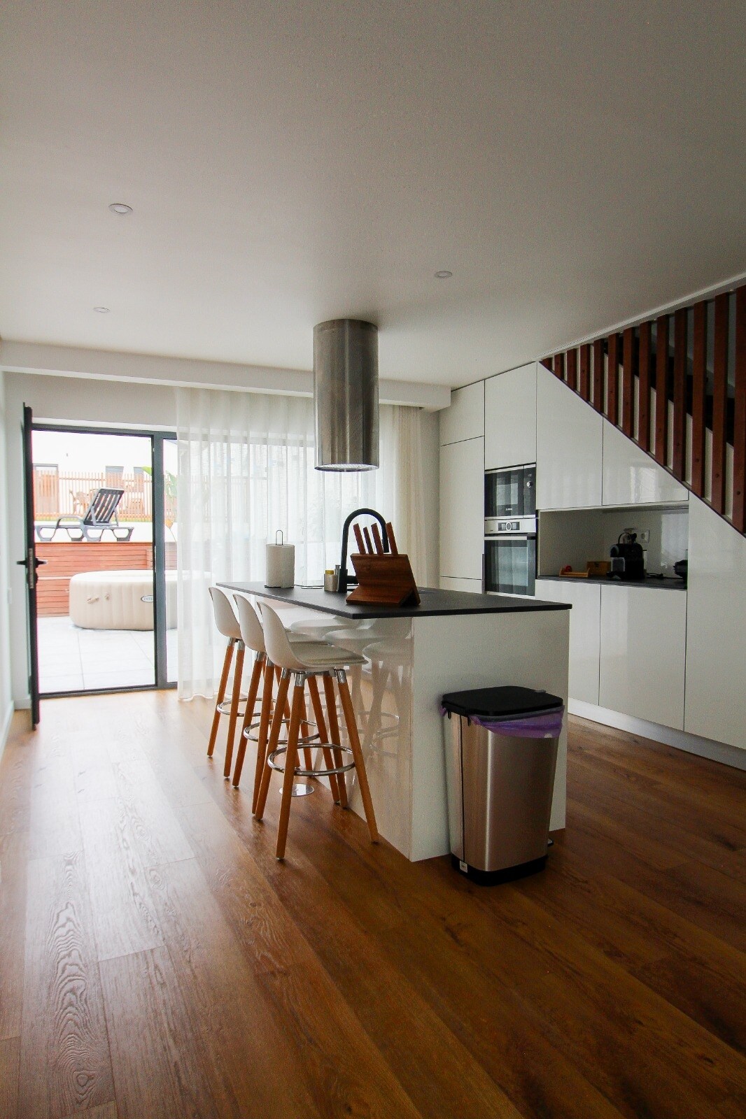 A modern kitchen area is seen featuring sleek white cabinetry and a central island with three wooden bar stools. Natural light flows in through large glass doors leading to a terrace. A stainless steel range hood hangs above the cooking area, while a kitchen appliance is visible nearby.