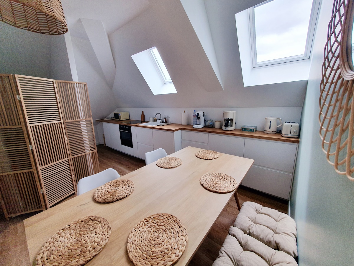 A modern kitchen area is displayed with slanted ceilings and skylights allowing natural light to fill the space. A wooden dining table is surrounded by white chairs, with woven mats placed on the table. Kitchen appliances, including a kettle and coffee maker, are neatly arranged on countertops.