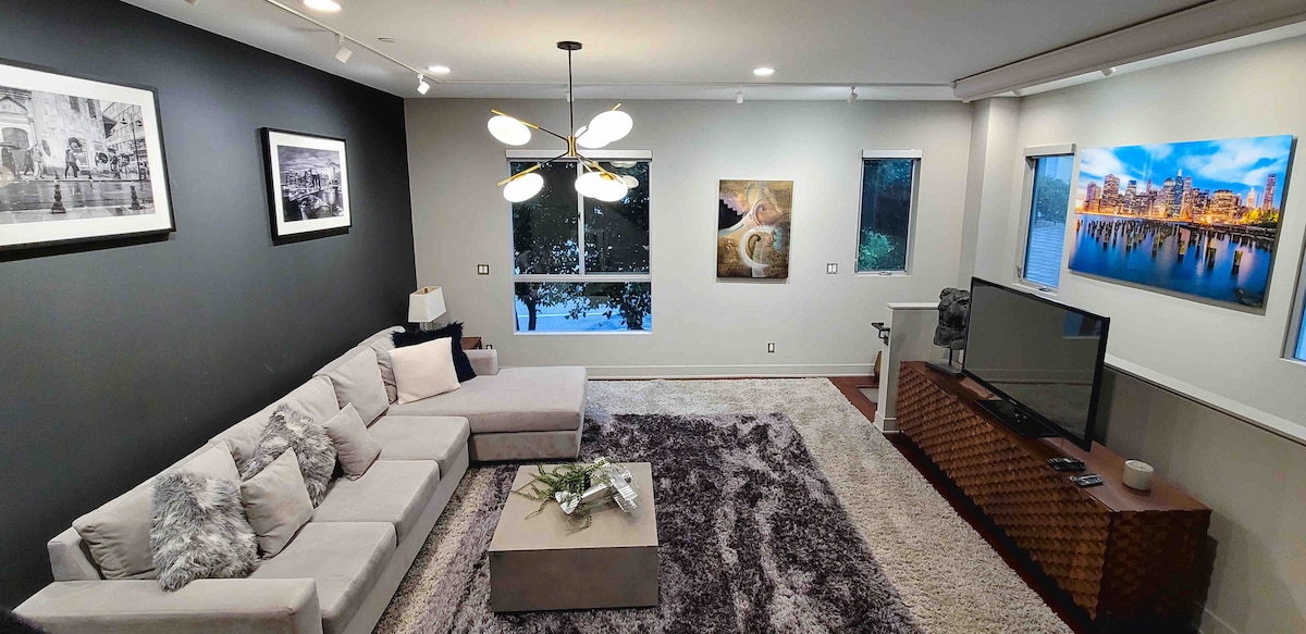 A spacious living area showcases a large sectional sofa, complemented by a plush area rug and tasteful decor. Two windows allow natural light to illuminate the room, while art pieces and a television enhance the ambiance of comfort and relaxation.