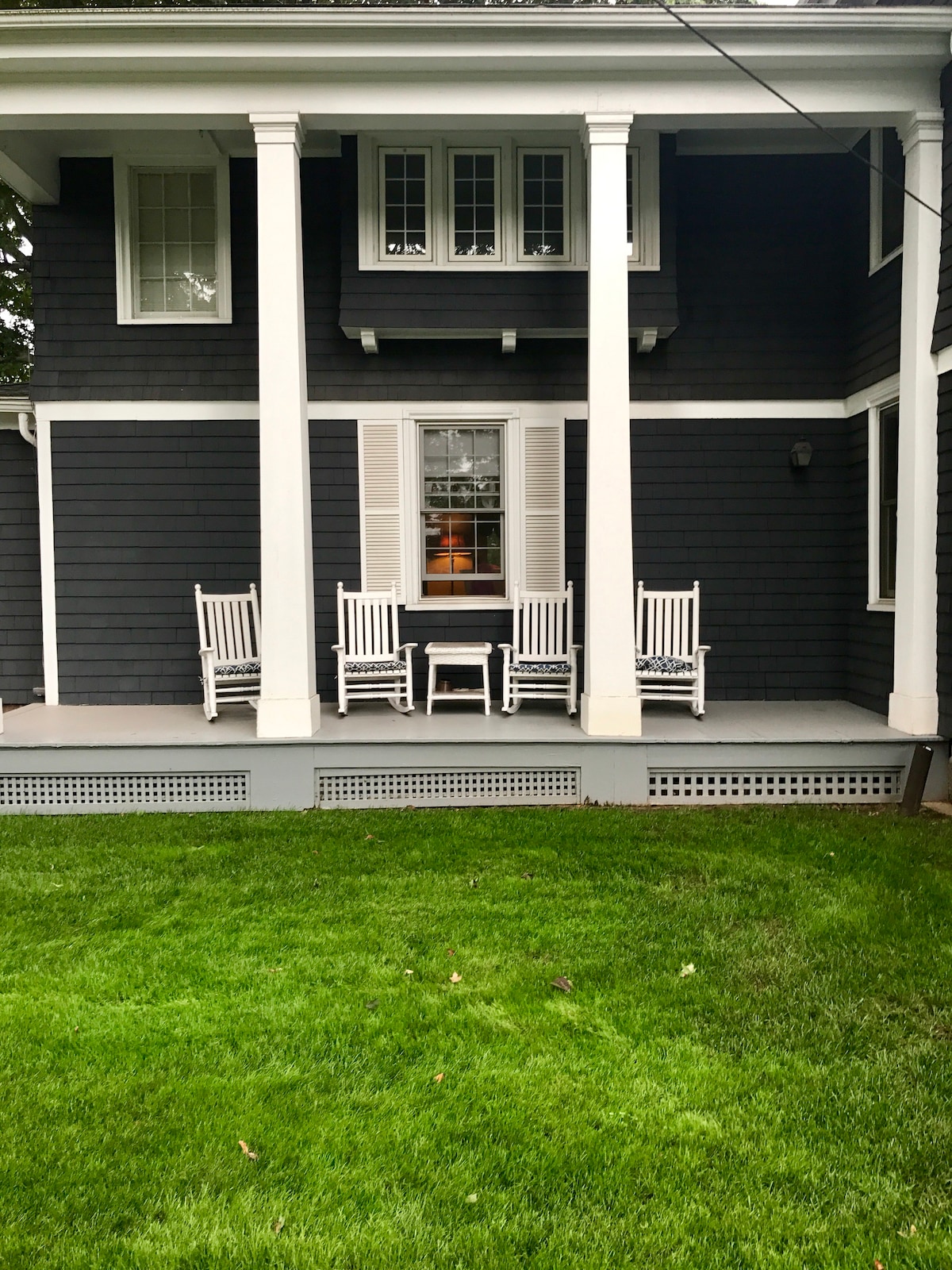 A welcoming porch area is showcased, featuring four white rocking chairs arranged on a covered deck. The dark exterior of the house is complemented by neutral shutters on the windows. A manicured lawn extends in front of the porch, providing a relaxing outdoor space.