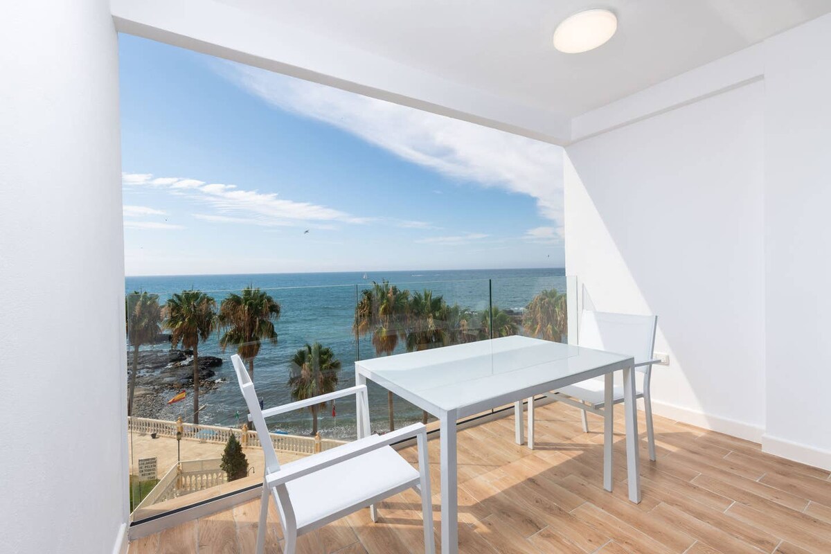 A glass table with two chairs overlooks the sea, providing a clear view of the coastline and palm trees. Natural light fills the space, highlighting the modern design and clean lines of the area. The scene evokes a sense of tranquility and connection to nature.