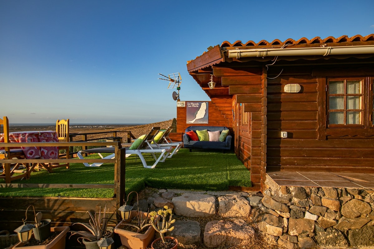 The charming cottage exterior features a wooden facade with a terracotta roof. A cozy sitting area is visible on the terrace, furnished with colorful cushions and flanked by sun loungers. Ocean views and an outdoor dining space enhance the inviting outdoor atmosphere.