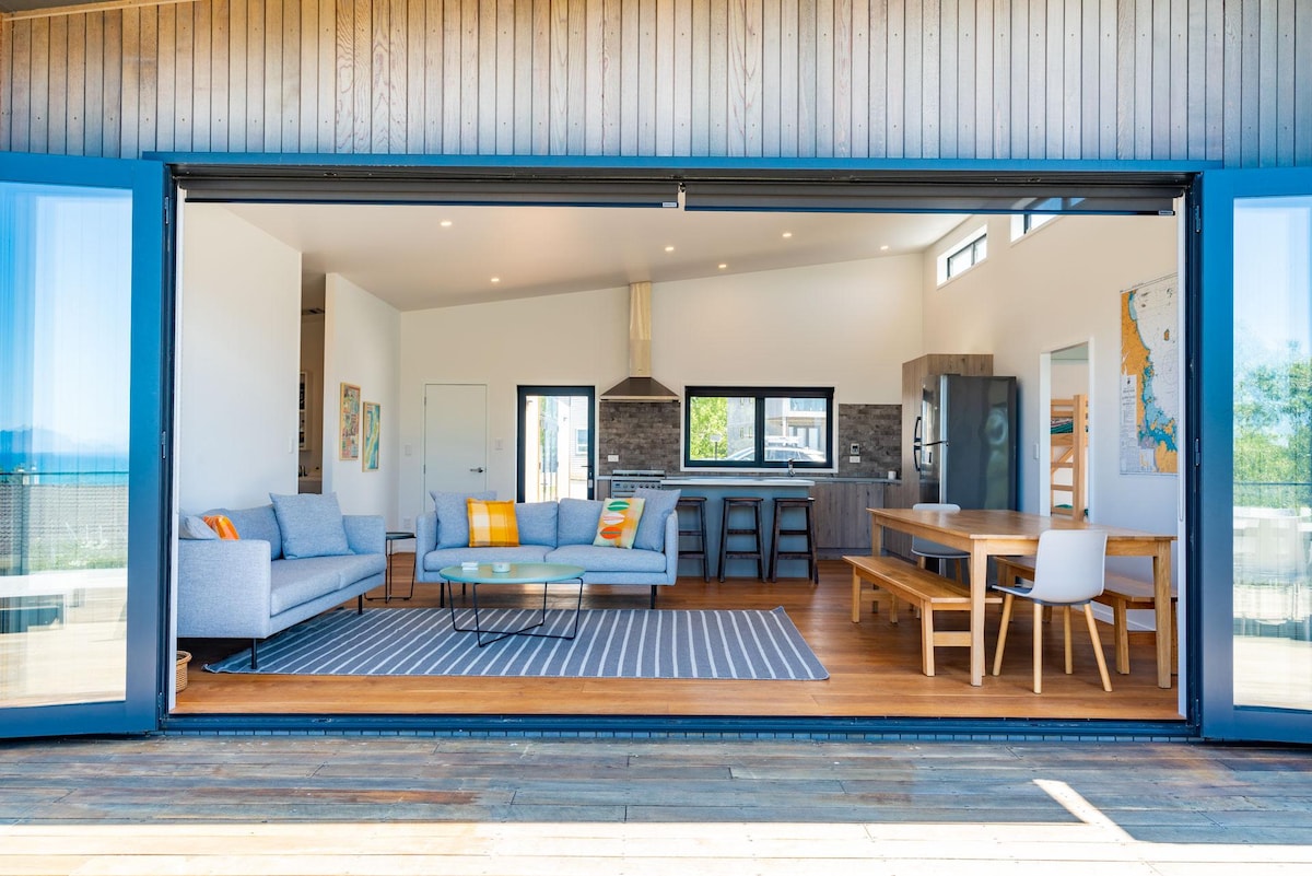An open-plan living area is showcased with large sliding doors providing a seamless transition to the deck. A comfortable seating arrangement includes a light gray couch and occasional chairs, accompanied by a modern dining table. Natural light enhances the relaxed and spacious atmosphere.