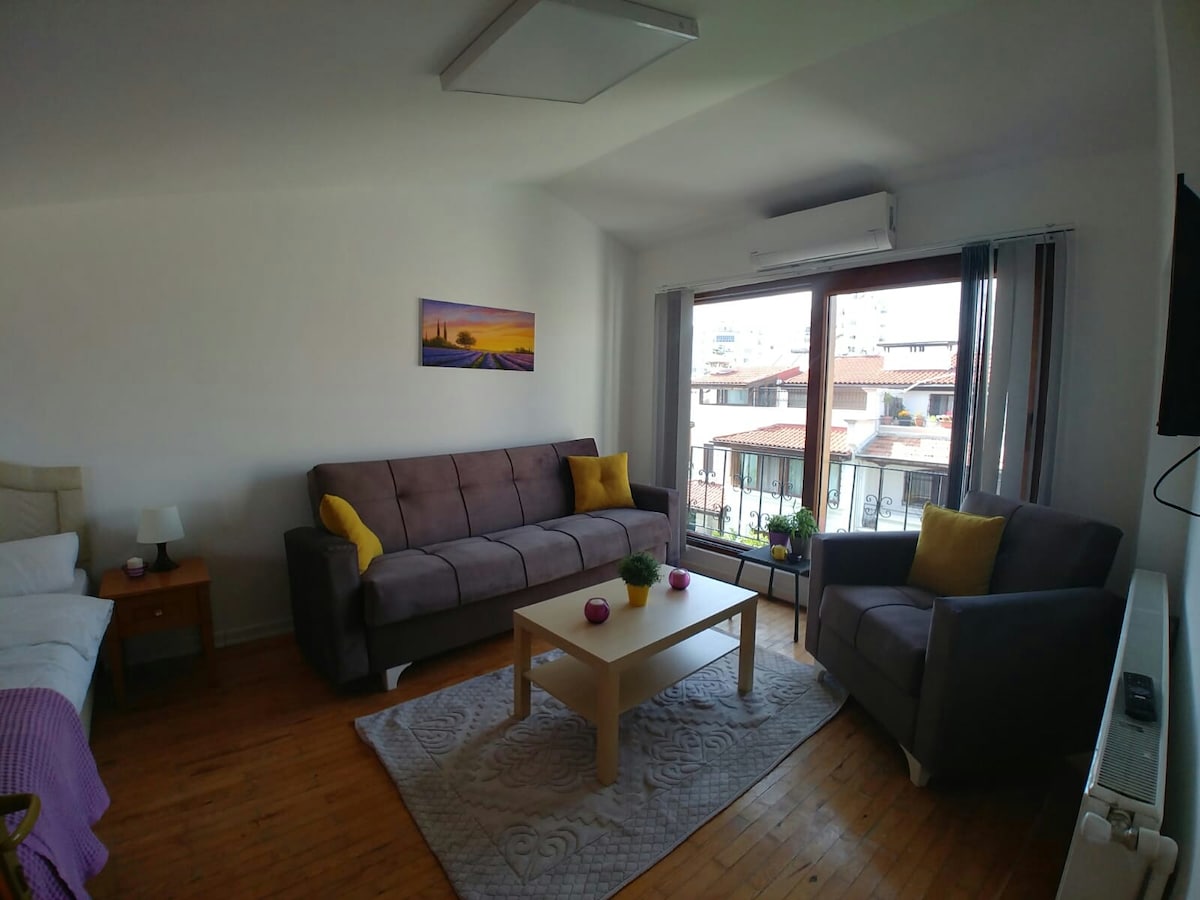 A living area features a gray sofa and armchair adorned with yellow cushions. A light wooden coffee table sits in the center, surrounded by small decorative items. Large windows allow natural light to fill the room, showcasing views of neighboring balconies.