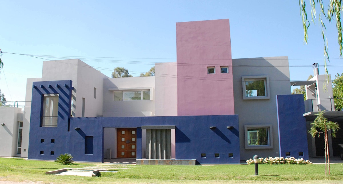 A modern home features a striking facade with a combination of colors, including purple and blue. The entrance is framed by geometric designs, and large windows allow natural light to illuminate the interior. Lush greenery and landscaping enhance the inviting exterior.