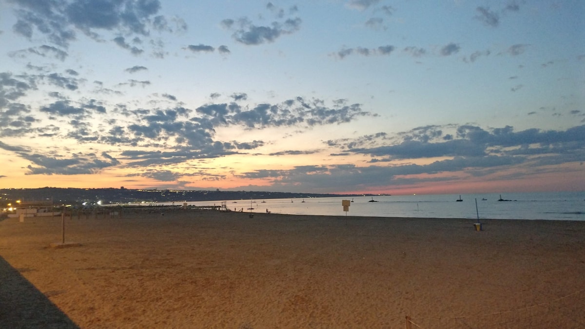 A wide sandy beach is seen at dusk, with the sky displaying a blend of soft colors. The horizon features silhouettes of boats, while distant lights from the nearby town twinkle. Gentle waves are visible against the shoreline, creating a peaceful coastal scene.