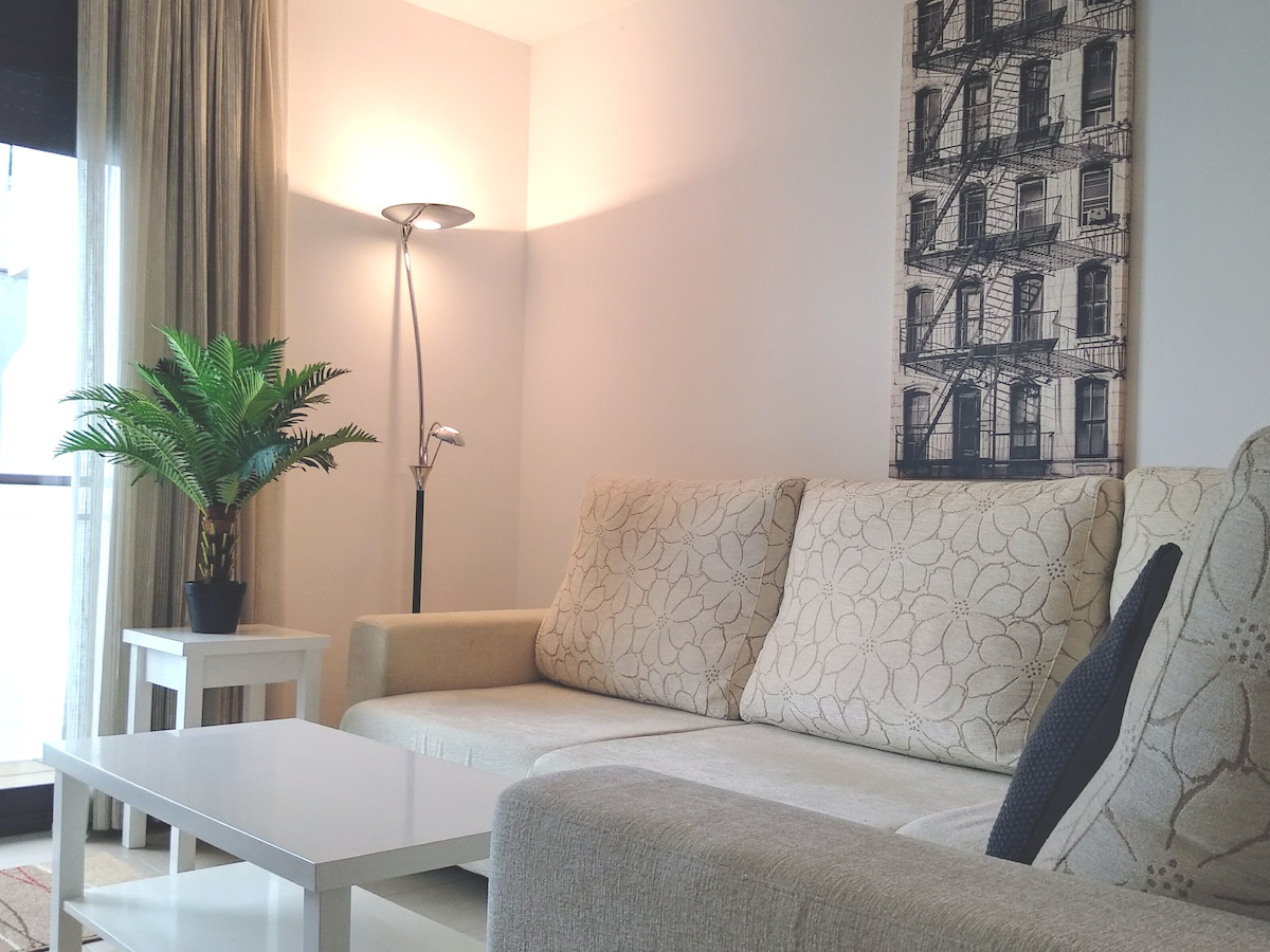 A comfortable seating area features a light-colored sofa adorned with subtle floral patterns. A modern floor lamp stands beside it, providing soft lighting. A small table is positioned in front of the sofa, complemented by a decorative green plant and a piece of framed wall art.