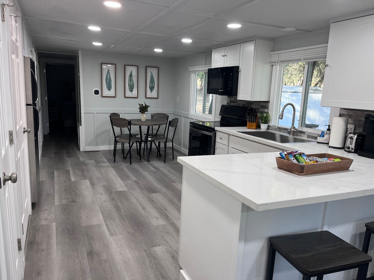A modern kitchen is shown with white cabinetry and a spacious island. A dining area features a round table with four chairs, surrounded by large windows. A tray of snacks is placed on the island, alongside essential appliances that enhance functionality.