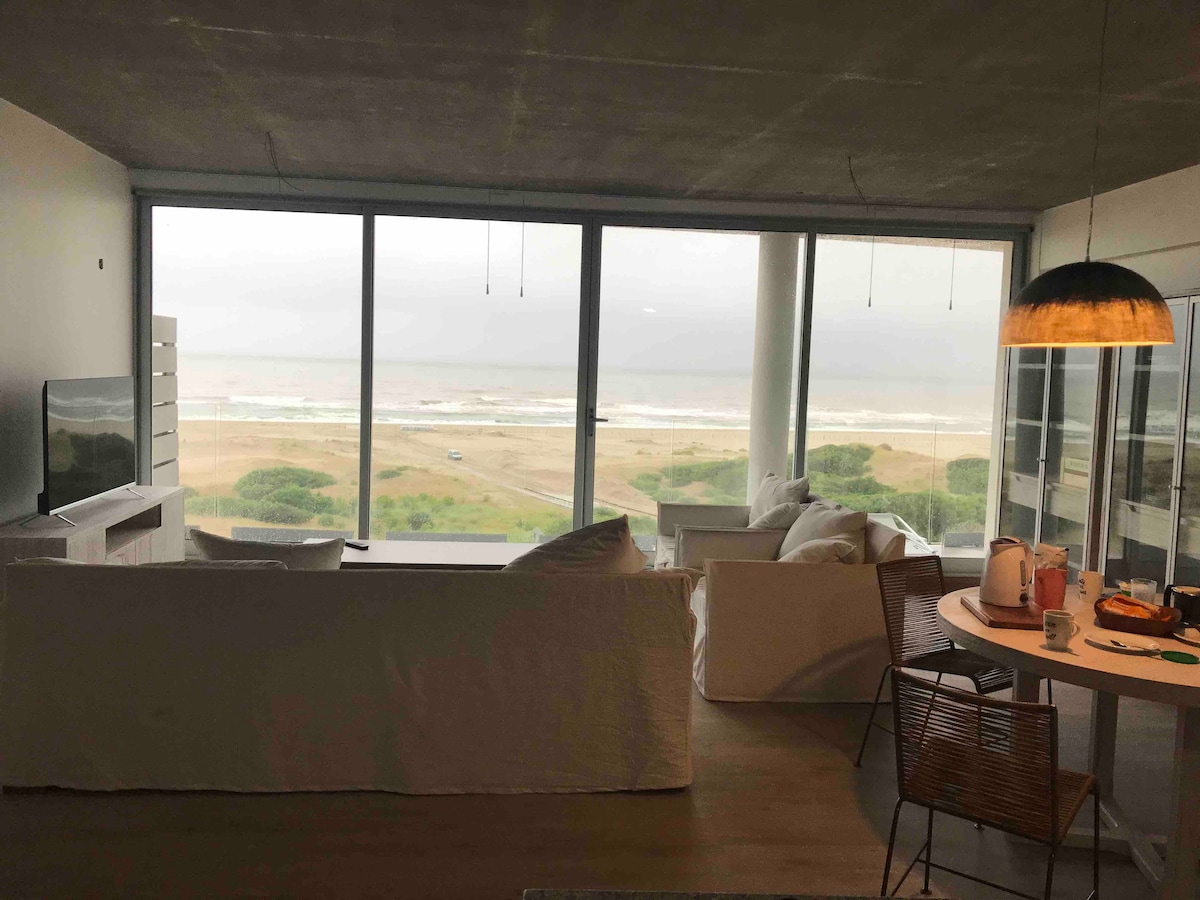 A spacious living area features large floor-to-ceiling windows overlooking the sea. A comfortable white sofa and modern dining table are arranged within the open space. Natural light is accentuated by neutral decor and simple furnishings, creating a welcoming environment.