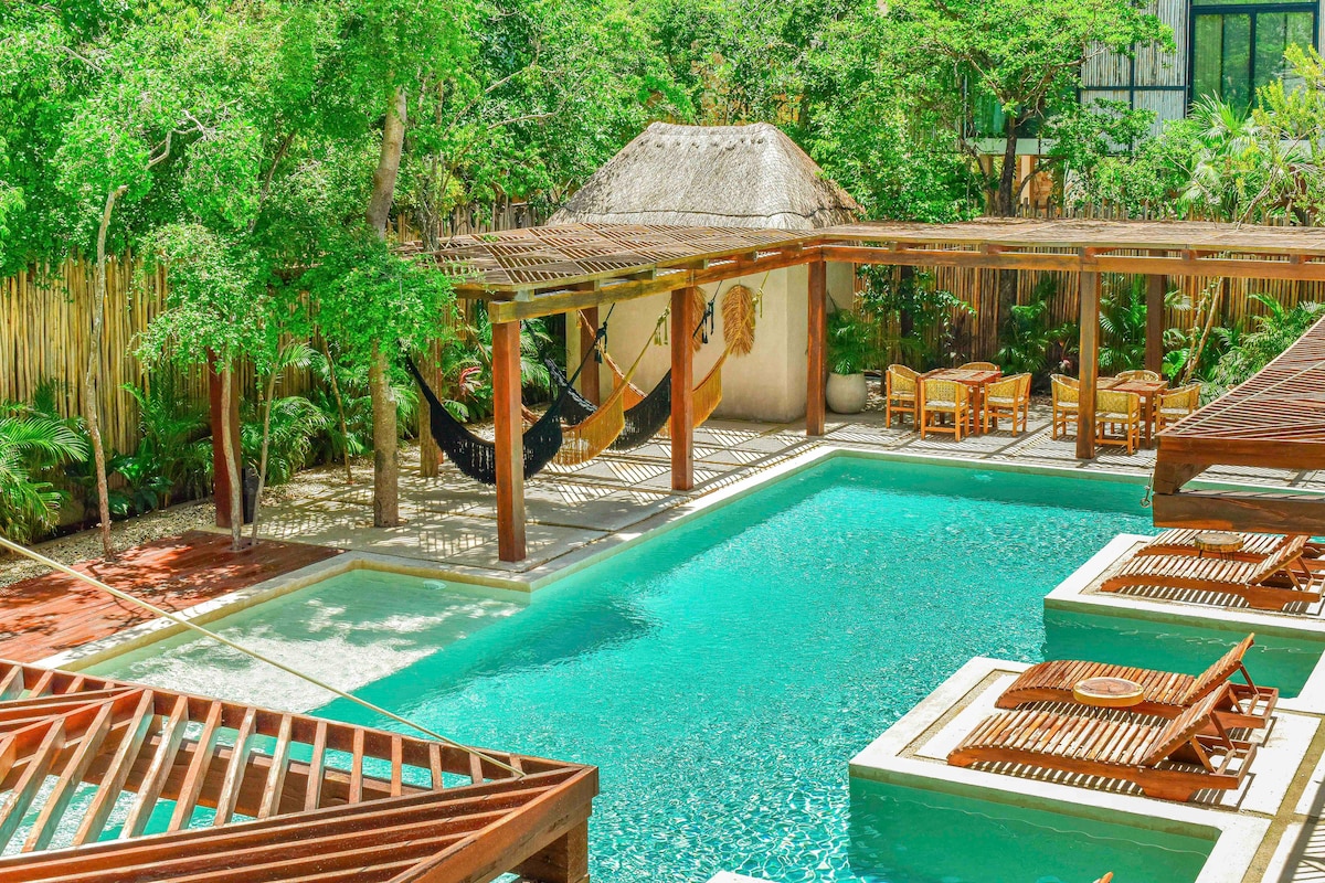 A large swimming pool is surrounded by lush greenery and wooden cabanas. Hammocks are draped under a thatched roof, providing a relaxed space. Sun loungers are positioned along the pool's edge, inviting guests to enjoy the tropical ambiance.