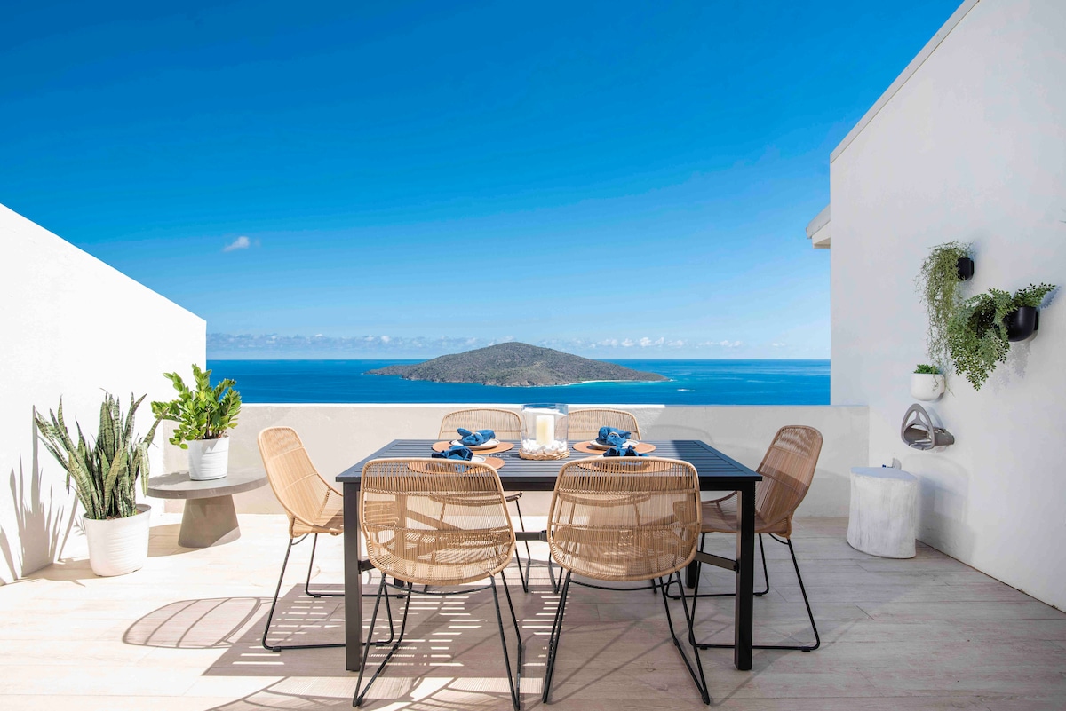 An inviting outdoor dining area features a black table surrounded by six wicker chairs. Lush greenery is present in a large pot nearby. The backdrop showcases panoramic ocean views under a clear blue sky, enhancing the tranquil atmosphere.