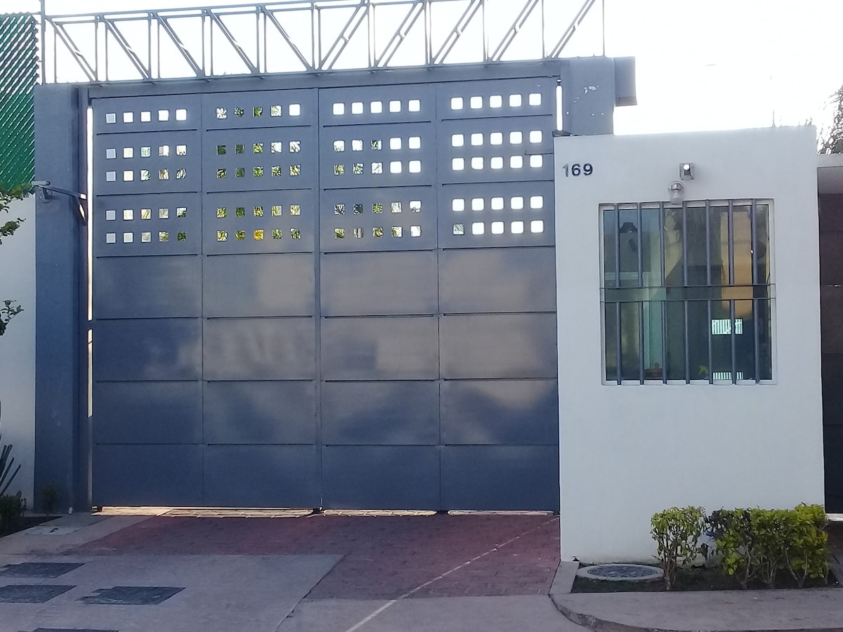 The image shows a modern gray gate with a grid pattern on the upper section, allowing for natural light. To the right, a white wall features a square window, providing a glimpse of the indoor space. Neatly trimmed shrubs border the entrance pathway.