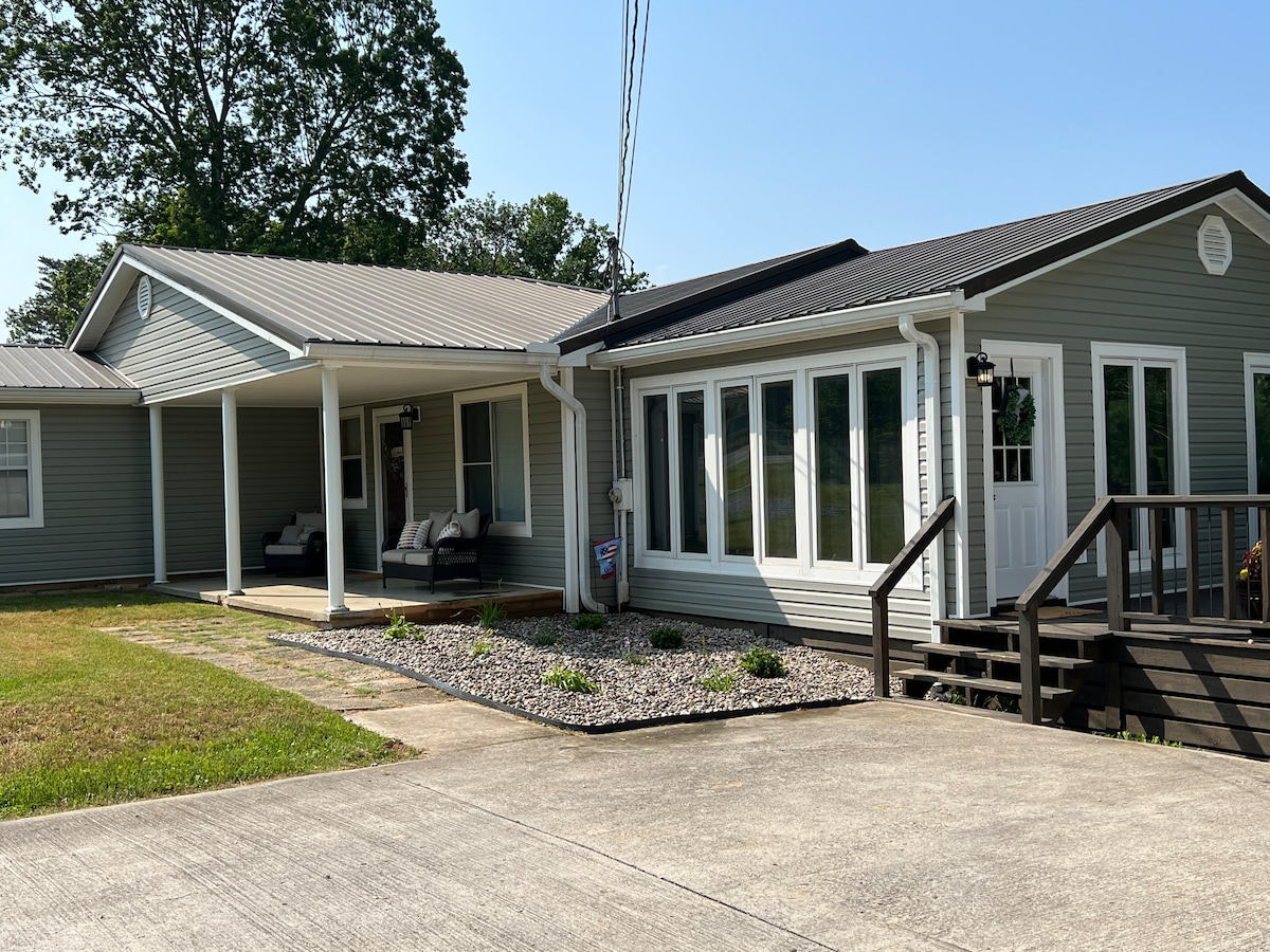 The exterior of the house features a spacious deck with seating arrangements, surrounded by a well-maintained lawn and gravel landscaping. Large windows allow for natural light, while a welcoming entrance is balanced by a slightly elevated porch leading into the home.